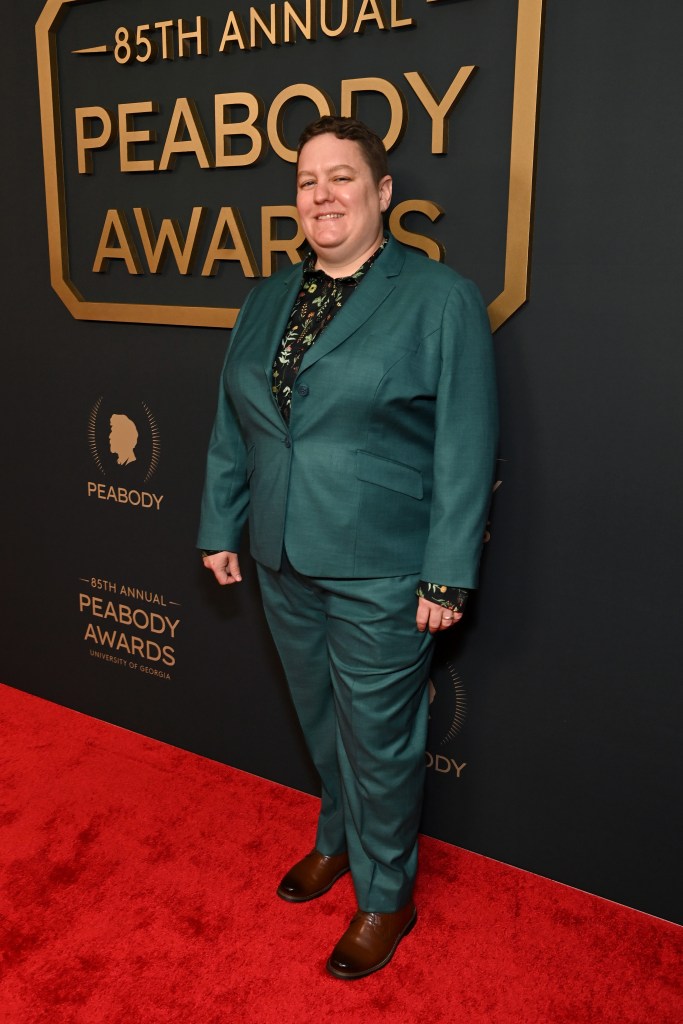 Adrienne Shaw, white woman with short hair, in a green suit stands on a red carpet at the 85th Annual Peabody Awards, smiling confidently against a black backdrop with gold lettering.
